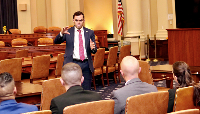 A man in a dark suit and pinkish necktie addresses a row of people in brown chairs.