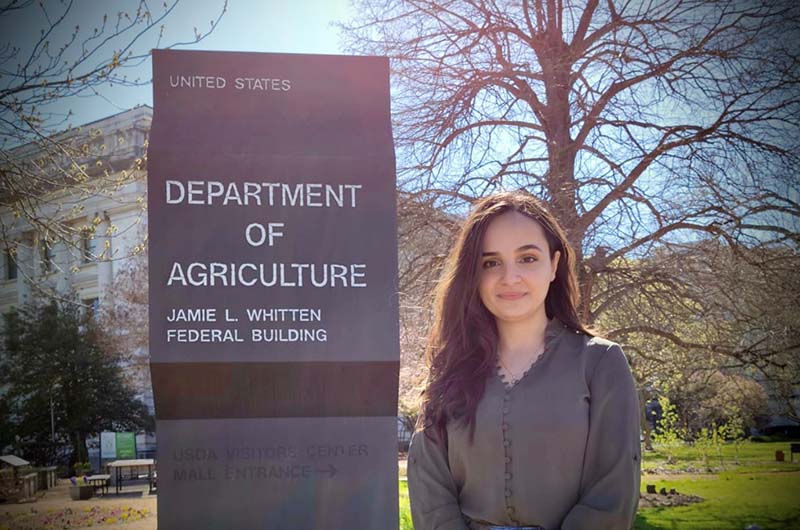 A young woman with brown hair poses in front of a federal building and its sign in Washington, D.C.