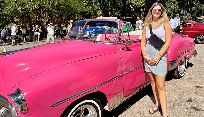 A young woman in a white outfit poses next to an antique pink car.