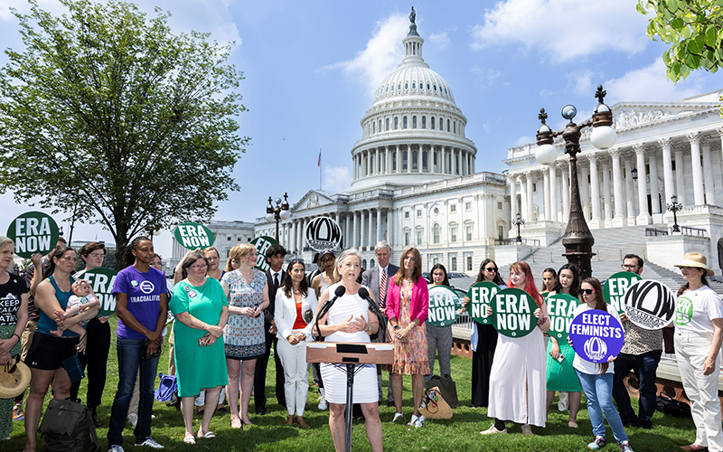 A woman in a white dress stands at a microphone in front of the U.S. Capitol while onlookers holding ERA Now signs surround her.