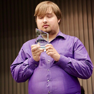 A blond man in a purple shirt gazes at a glass trophy in his hands.