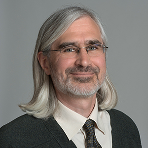 A man in eyeglasses with long silver hair to his shoulders wears a black tie and a white shirt.