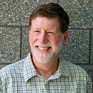 A man in a green checked shirt with brown hair and a short white beard smiles off camera.