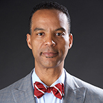 Michael Fauntroy wearing a gray suit, blue shirt, and red bow tie with yellow stripes.