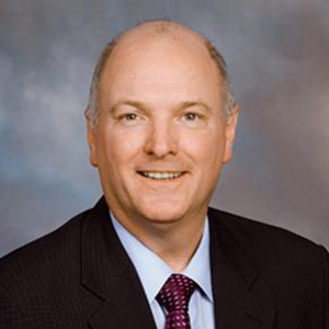 Terry Clower standing and smiling while wearing a dark suit, blue collared shirt and a tie