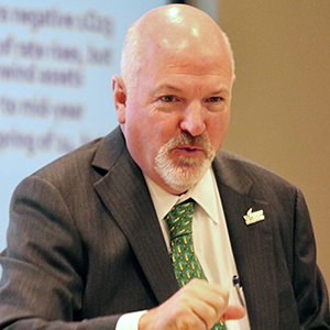 A bald man with a short white beard in a green necktie stands in front of a PowerPoint screen.