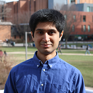 Mahid Sheikh stands outside on the George Mason University Fairfax Campus while wearing a blue collared shirt and smiling.