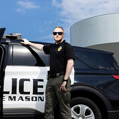 A man in a police uniform and wearing sunglasses stands next to a police car with the Mason water tower behind it.