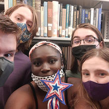 Five students in masks stand in front of bookshelves.