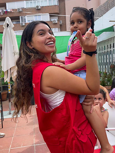 A young woman in a red vest and with long dark hair holds a toddler with an ice cream in her mouth.