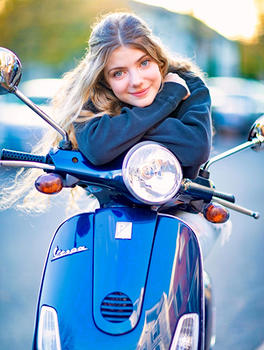 A young woman on a blue scooter gazes at the camera over the handlebars.