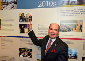 James P. Danoy wearing a dark suit and a red necktie points to a photo of himself with President Obama on a wall.
