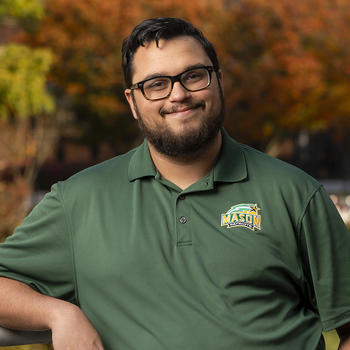 Zayd Hamid stands outside while smiling and wearing a green George Mason University collared shirt